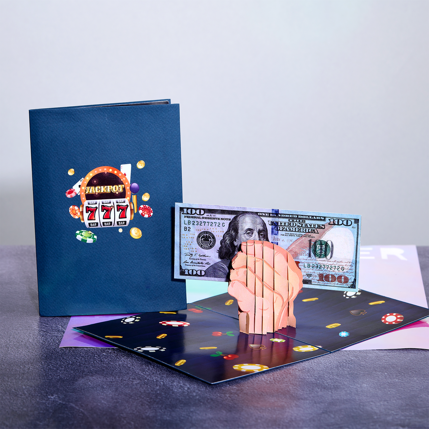 An open 3D pop-up card showing a clenched fist holding a $100 bill, with poker chips and casino-themed decorations. Next to it is a closed blue card featuring a slot machine illustration with Jackpot text
