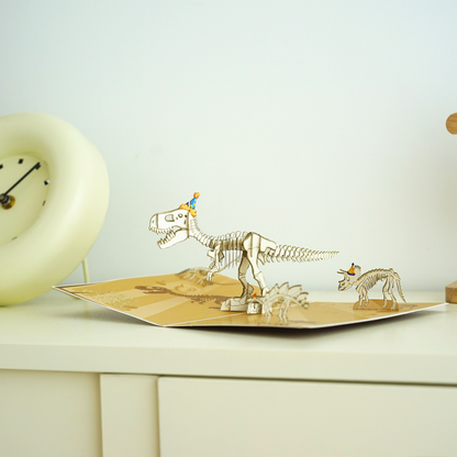 A pop-up card featuring a T-Rex skeleton wearing a party hat, standing on a beige base with fossil illustrations. A smaller dinosaur skeleton, also with a party hat, is positioned nearby. A clock is in the background.