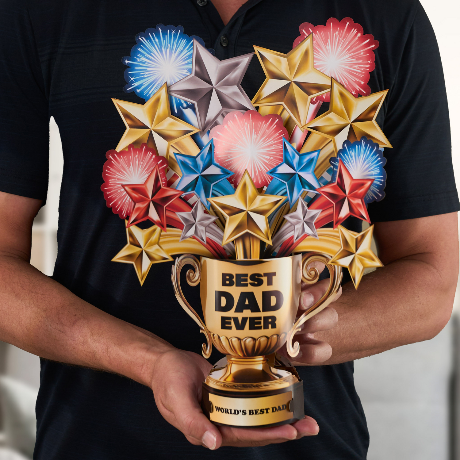 A person in a black shirt holds a 3D pop-up greeting card shaped like a golden trophy. The trophy features the words "BEST DAD EVER" on the front and "WORLD'S BEST DAD" on the base. Colorful stars and fireworks in red, blue, gold, and silver burst out from the top of the trophy, creating a festive and celebratory look.