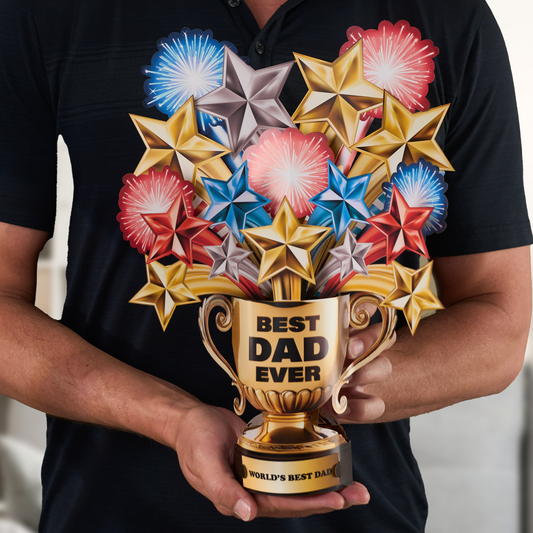 A person in a black shirt holds a 3D pop-up greeting card shaped like a golden trophy. The trophy features the words "BEST DAD EVER" on the front and "WORLD'S BEST DAD" on the base. Colorful stars and fireworks in red, blue, gold, and silver burst out from the top of the trophy, creating a festive and celebratory look.