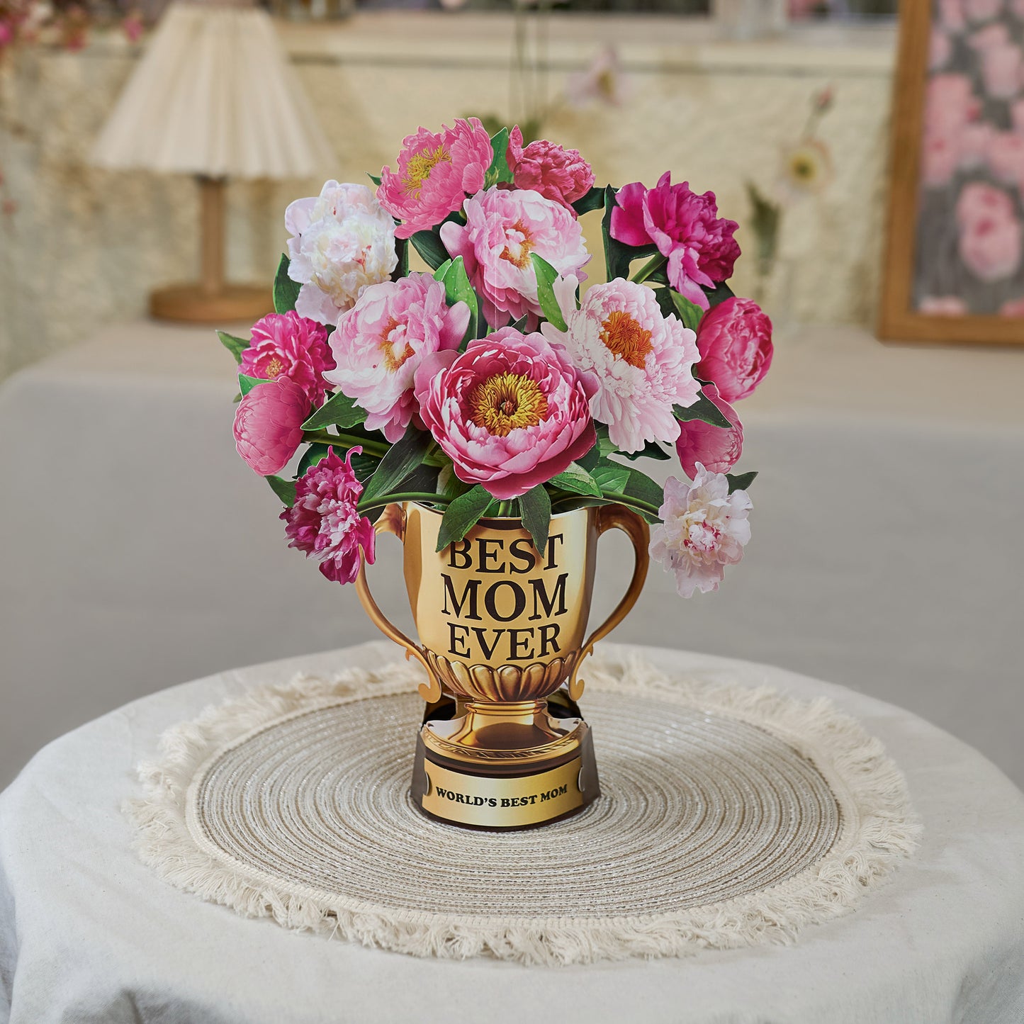 A pop-up Mother's Day card displayed on a round woven placemat atop a neutral-toned table. The card features a golden trophy labeled "BEST MOM EVER" filled with vibrant pink and white peonies. The base of the trophy reads "WORLD'S BEST MOM." The cozy, home-like setting in the background includes soft lighting, a table lamp, and floral decor, creating a warm and heartfelt atmosphere, perfect for celebrating a special mom.