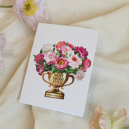 A folded greeting card placed on a soft, neutral fabric background, surrounded by delicate pink and white flower petals. The card's front features a golden trophy labeled "BEST MOM EVER" filled with blooming pink and white peonies. The base of the trophy reads "WORLD'S BEST MOM." The floral and cozy presentation highlights a heartfelt and celebratory message, perfect for Mother's Day or expressing appreciation for a special mom.