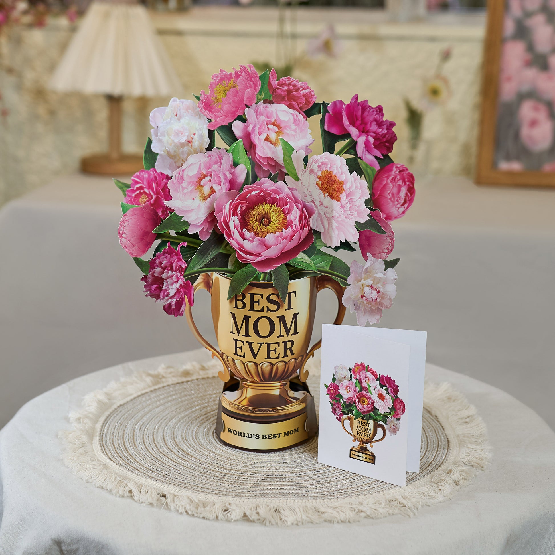 A Mother's Day pop-up card displayed on a round woven placemat atop a neutral-toned table. The card features a golden trophy labeled "BEST MOM EVER" filled with vibrant pink and white peonies. The base of the trophy reads "WORLD'S BEST MOM." Next to the pop-up card is a smaller folded greeting card with the same trophy and floral design. The cozy, homey setting in the background includes soft lighting, a table lamp, and floral decor, emphasizing a warm and heartfelt ambiance.