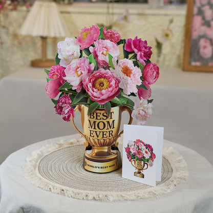 A Mother's Day pop-up card displayed on a round woven placemat atop a neutral-toned table. The card features a golden trophy labeled "BEST MOM EVER" filled with vibrant pink and white peonies. The base of the trophy reads "WORLD'S BEST MOM." Next to the pop-up card is a smaller folded greeting card with the same trophy and floral design. The cozy, homey setting in the background includes soft lighting, a table lamp, and floral decor, emphasizing a warm and heartfelt ambiance.