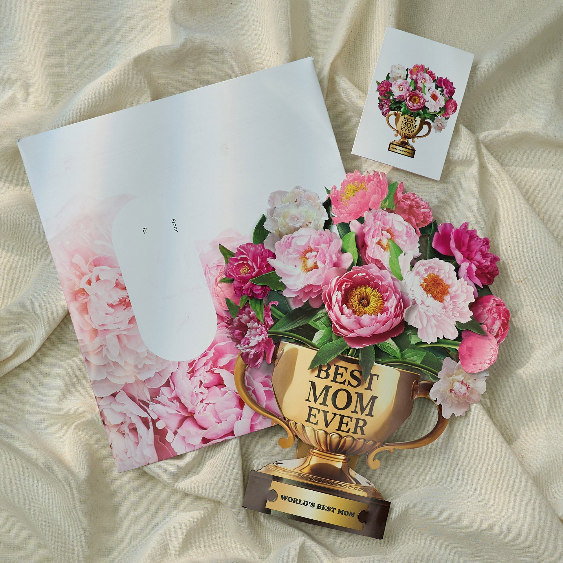 A Mother's Day greeting card set displayed on a soft fabric background. The set includes a pop-up card shaped like a golden trophy labeled "BEST MOM EVER" with vibrant pink and white peonies blooming from the top. The base of the trophy reads "WORLD'S BEST MOM." Next to the card is a floral-themed envelope with a designated "From" and "To" area, decorated with soft pink peonies. A matching mini card with the trophy and flowers is also shown, highlighting the thoughtful and celebratory nature of the gift.