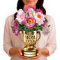 A woman holding a golden trophy-shaped vase labeled "BEST MOM EVER" with vibrant pink and white peonies blooming from the top. The base of the trophy reads "WORLD'S BEST MOM." The flowers are lush and colorful, conveying a celebratory and heartfelt message.
