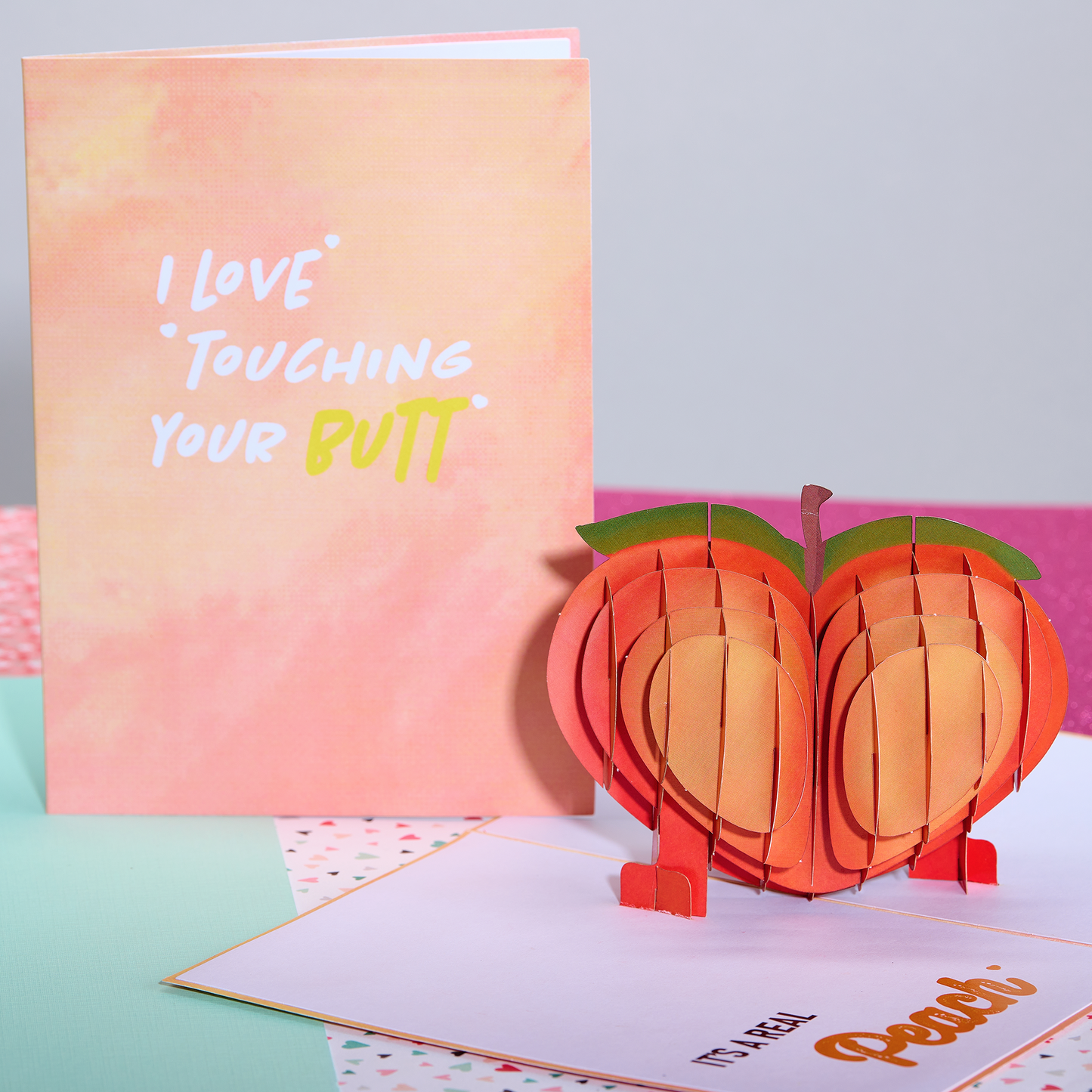 A pop-up card with a 3D peach shape, accompanied by a cover that reads I Love Touching Your Butt in playful white and yellow text on a pink and orange gradient background. Inside, the card says It's a real peach.