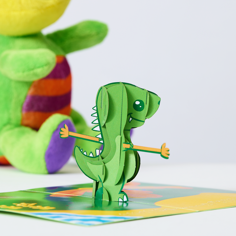 A green dinosaur on a green board, with a butterfly fluttering nearby. A delightful valentine's day card showcasing a pop-up T-rex with extended arms, and the heartfelt message 'always and forever' written in sand on a sandy beach.