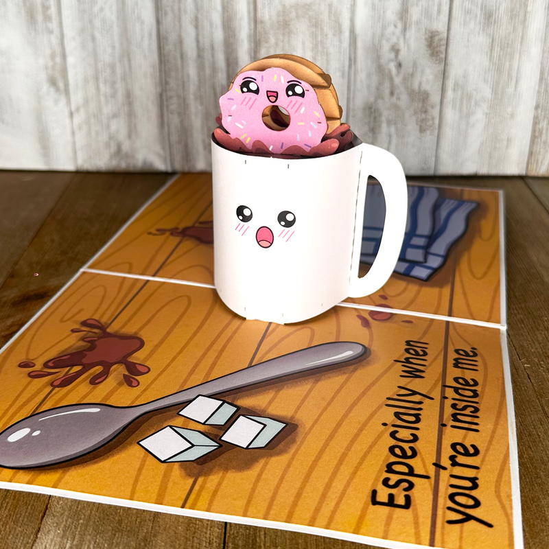  A cup with a donut inside, a spoon on a table. A humorous anniversary pop-up greeting card of a donut being dipped in coffee.