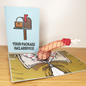 A pop-up card with a 3D pink and red worm ( DICK ) -like figure sits next to a blue card featuring an illustration of a mailbox with a package inside. The text on the blue card reads YOUR PACKAGE HAS ARRIVED!