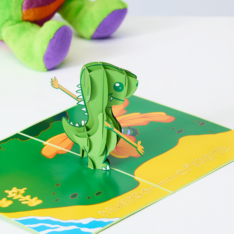 A green dinosaur on a green board with a butterfly. A cute valentine's day card with a pop-up T-rex and arm extenders, saying 'always and forever' on a sandy beach