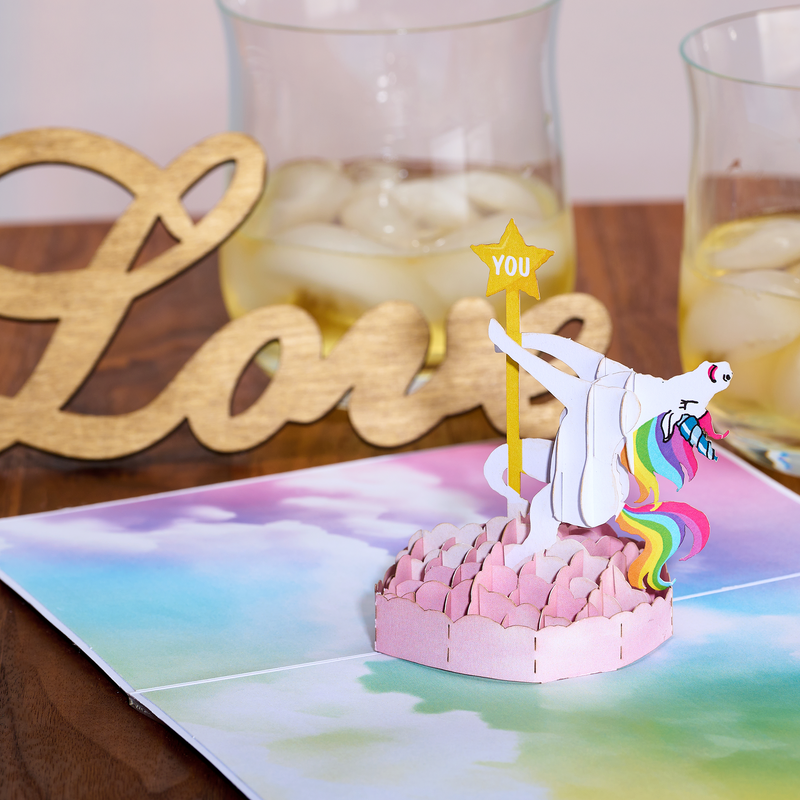 Funny pop up birthday card with a unicorn dancing on a pole, surrounded by clouds on a rainbow background.