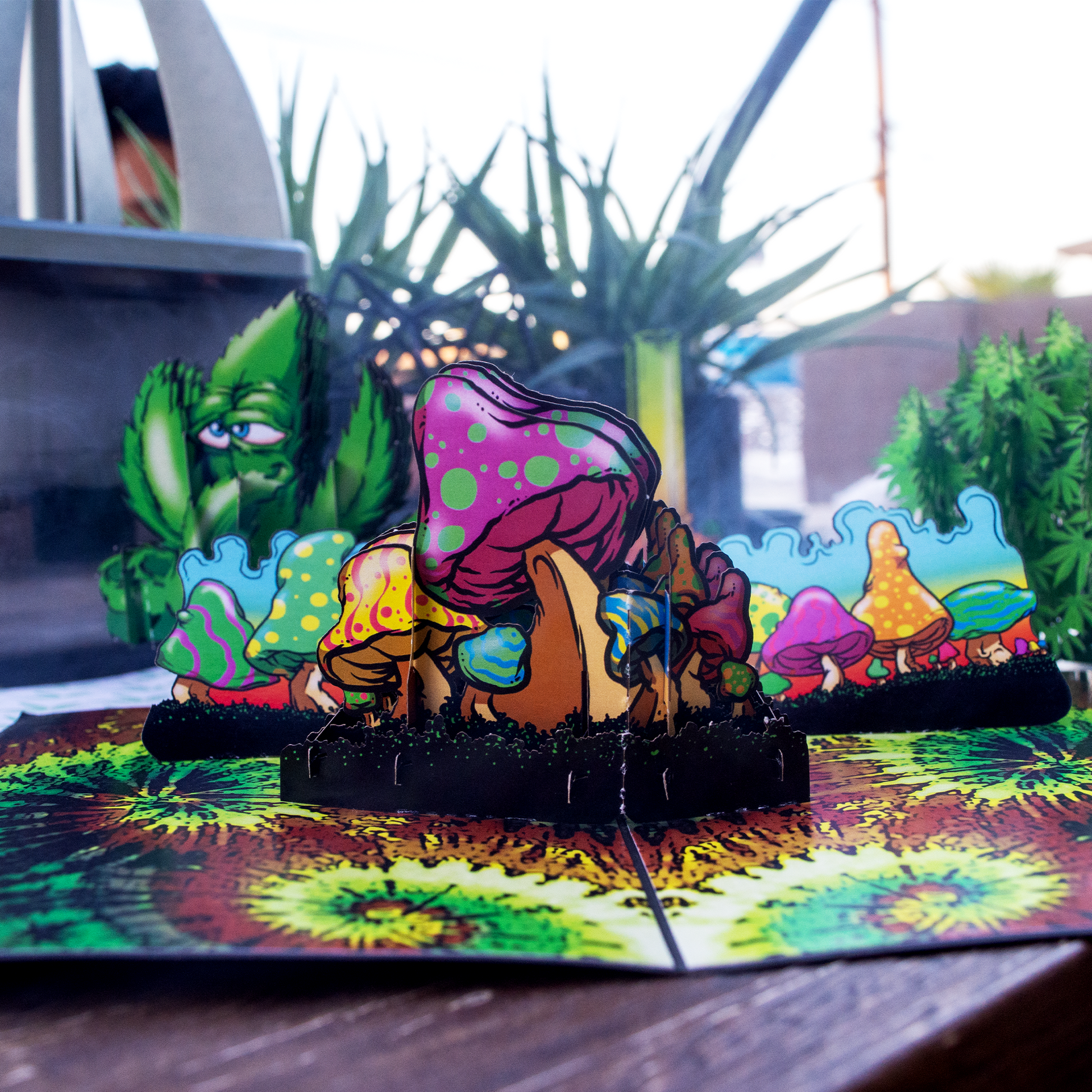 A pop-up card featuring vibrant, psychedelic mushrooms with colorful polka dots and swirls, set against a dynamic background with green plants and a whimsical, cartoonish cactus character in the distance.