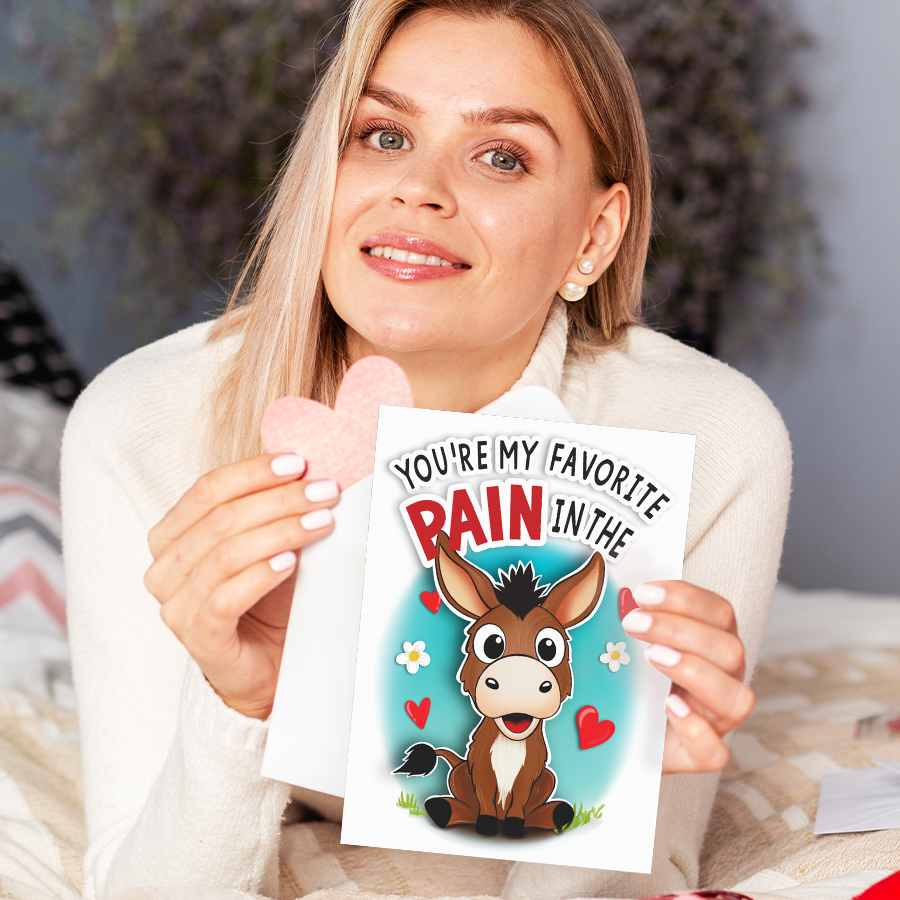 Smiling woman holding a greeting card featuring a cute donkey with hearts and the text "You're My Favorite Pain."