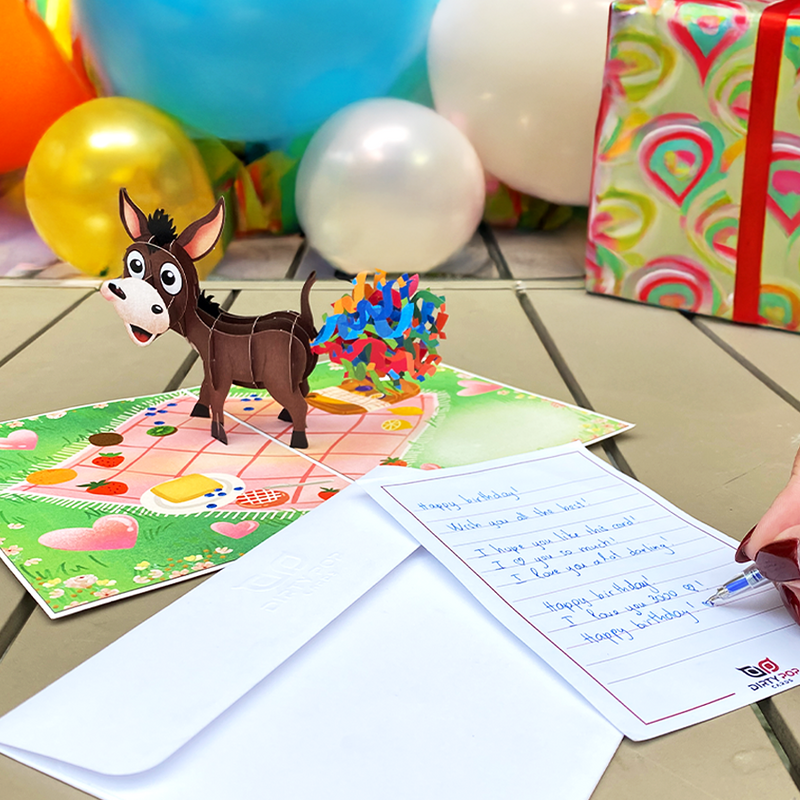 3D pop-up card featuring a donkey with confetti, alongside a handwritten note, envelope, balloons, and gifts.