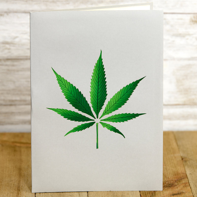 A greeting card with a green cannabis leaf illustration on a plain white background. The card is standing upright on a wooden surface, highlighting the simplicity and bold design of the leaf.