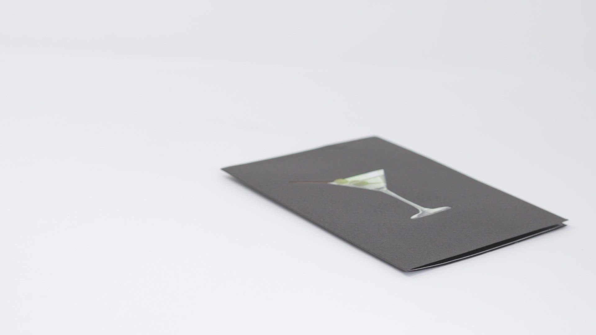 A video shows a pop-up card opening to reveal a 3D woman lounging in a martini glass, playfully holding a stirrer. The cover features a martini with olives, creating a fun and celebratory scene.