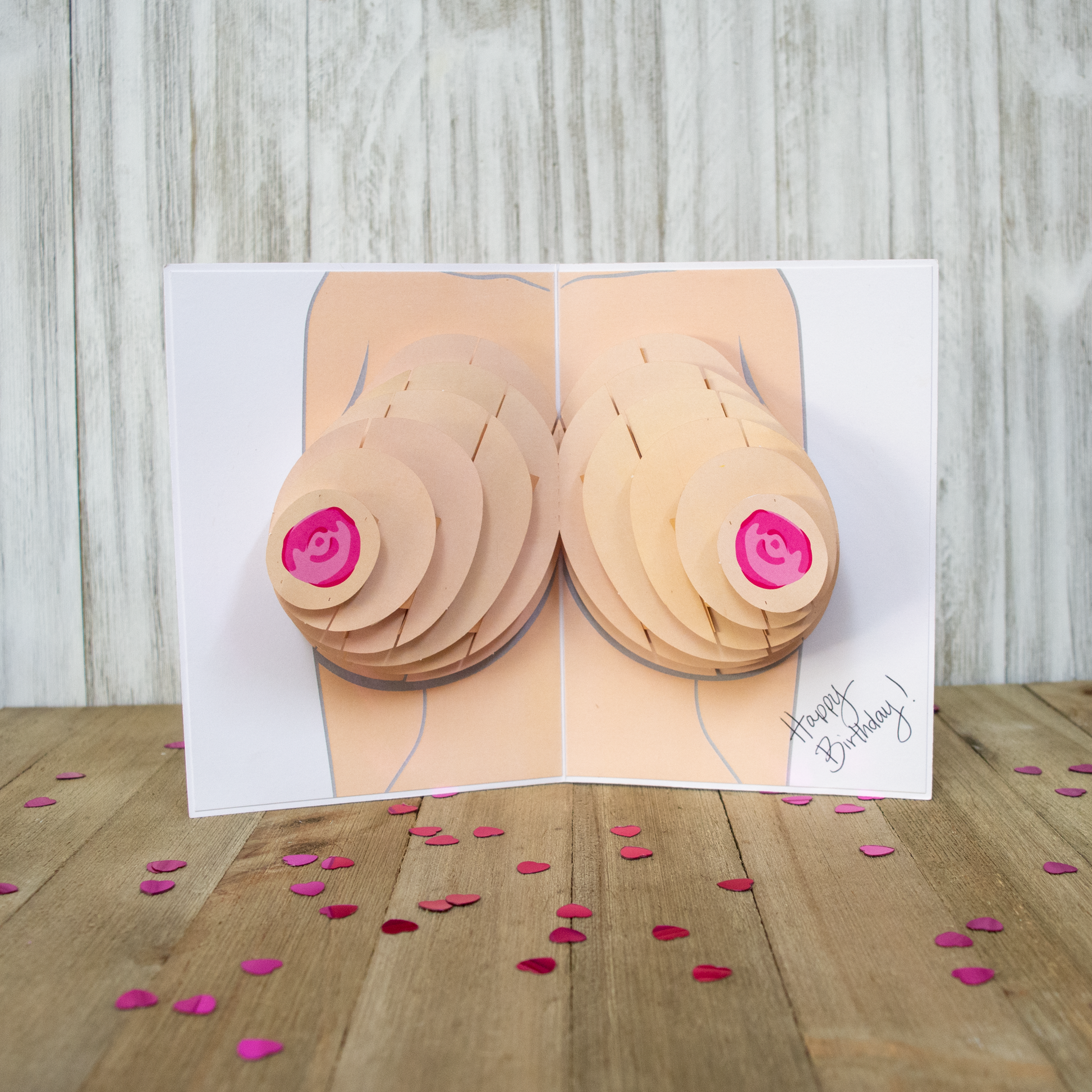 The image shows an open pop-up card with a 3D illustration of breasts, featuring layered paper elements and pink nipples, displayed on a wooden surface. Pink heart-shaped confetti is scattered around the card, which reads Happy Birthday!