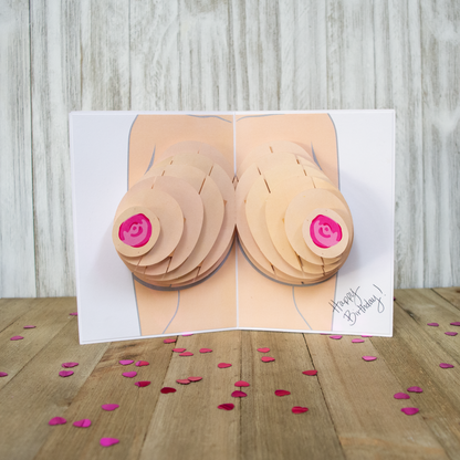 The image shows an open pop-up card with a 3D illustration of breasts, featuring layered paper elements and pink nipples, displayed on a wooden surface. Pink heart-shaped confetti is scattered around the card, which reads Happy Birthday!