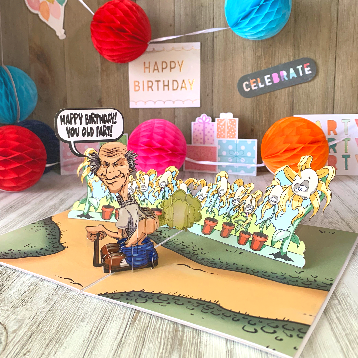 A playful pop-up birthday card shows a cartoon of an elderly man with a speech bubble saying HAPPY BIRTHDAY! YOU OLD FART! set against a colorful background with potted plants and festive decorations, including balloons and signs.