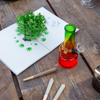 A pop-up card displaying a 3D cannabis plant lies open on a wooden table next to a colorful glass bong and three rolled joints. The setting suggests a relaxed, recreational atmosphere.