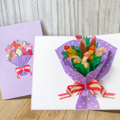 An open 3D pop-up card with a purple bouquet featuring stylized shapes and a red and yellow ribbon, next to a closed card with a similar colorful bouquet illustration on a wooden surface