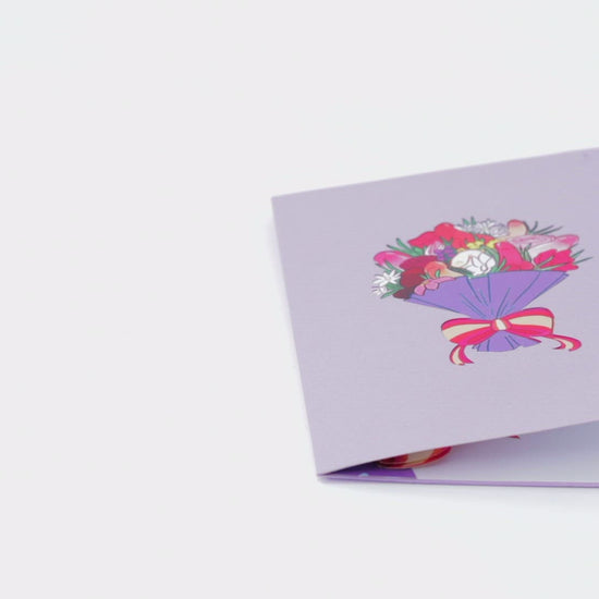 Video of a purple greeting card with a colorful bouquet illustration. The card is opened to reveal a 3D pop-up bouquet with vibrant, stylized flowers tied with a red and yellow ribbon