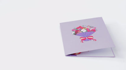 Video of a purple greeting card with a colorful bouquet illustration. The card is opened to reveal a 3D pop-up bouquet with vibrant, stylized flowers tied with a red and yellow ribbon