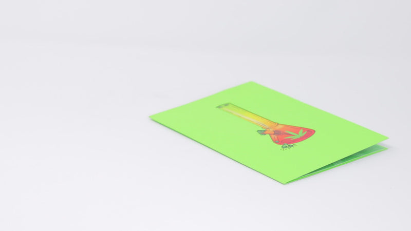 A video shows a pop-up card with a 3D bong design being opened and displayed. The camera pans around to capture the colorful details of the bong illustration, while a real bong and lighter are placed nearby, enhancing the playful theme.